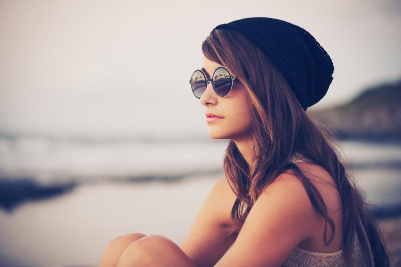 Žena v čapici a slnečných okuliaroch sediaca na pláži a pozerajúca do diaľky